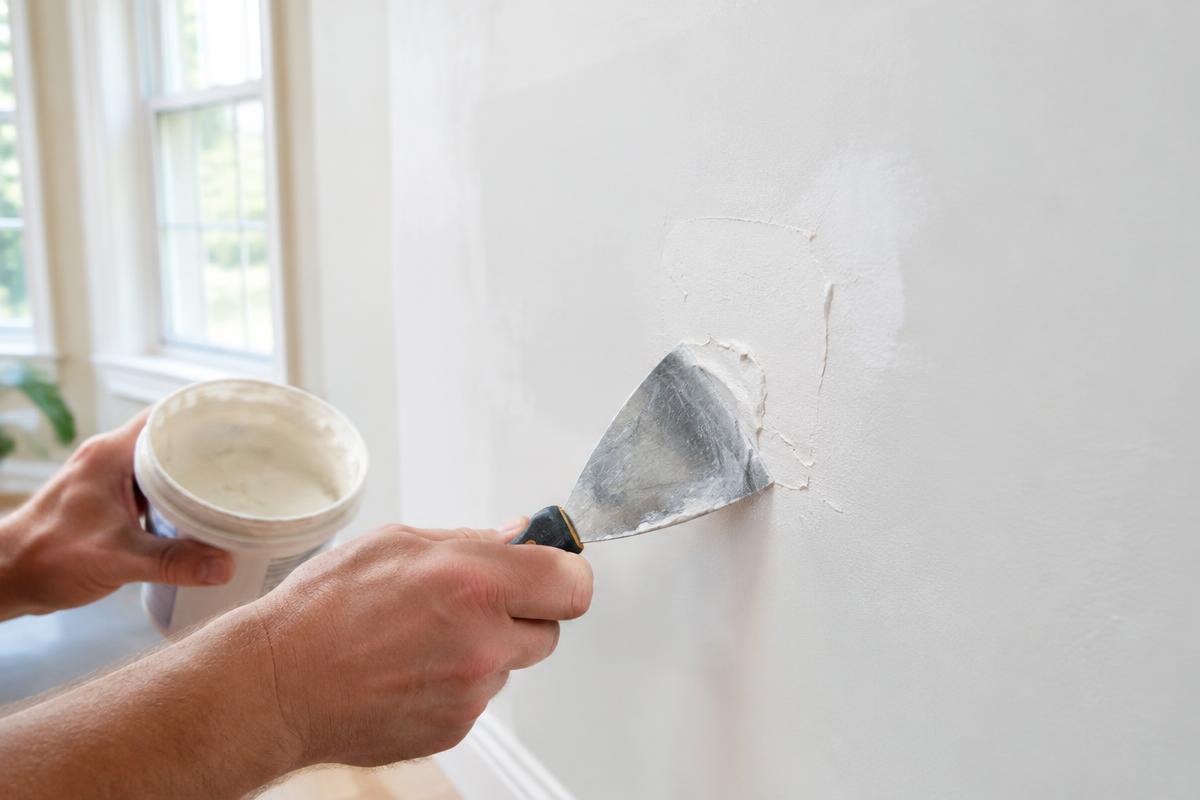 Hands patching a hole in a white drywall wall with a putty knife and spackle